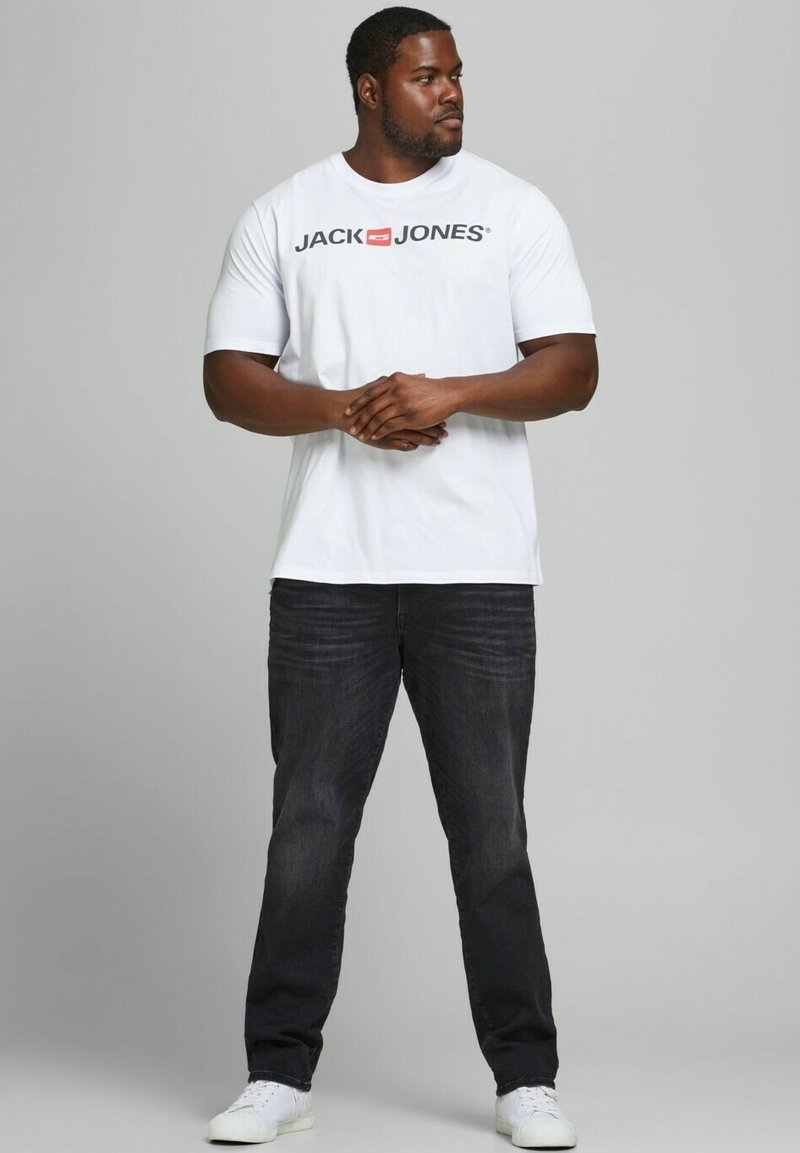 Weißes T-Shirt mit kurzen Ärmeln und dem "JACK & JONES"-Logo in Schwarz und Rot; getragen mit dunklen Jeans und weißen Sneakers. Modell steht mit verschränkten Armen.