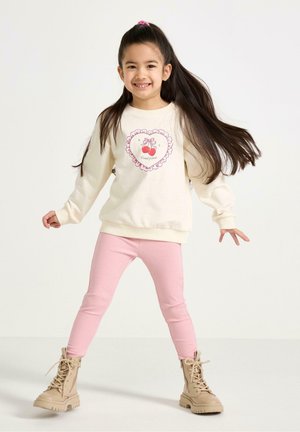Smilende pige med langt hår iført en cremefarvet sweatshirt med kirsebær, lyserøde leggings og lyse snørestøvler, stående foran en hvid baggrund.
