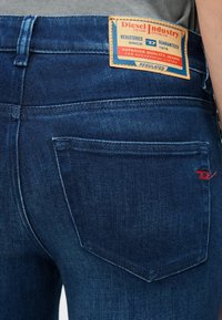 Tylnia kieszeń ciemnoniebieskich jeansów Diesel z beżowo-czerwoną metką z logo oraz subtelnym czerwonym przeszyciem na krawędzi kieszeni.