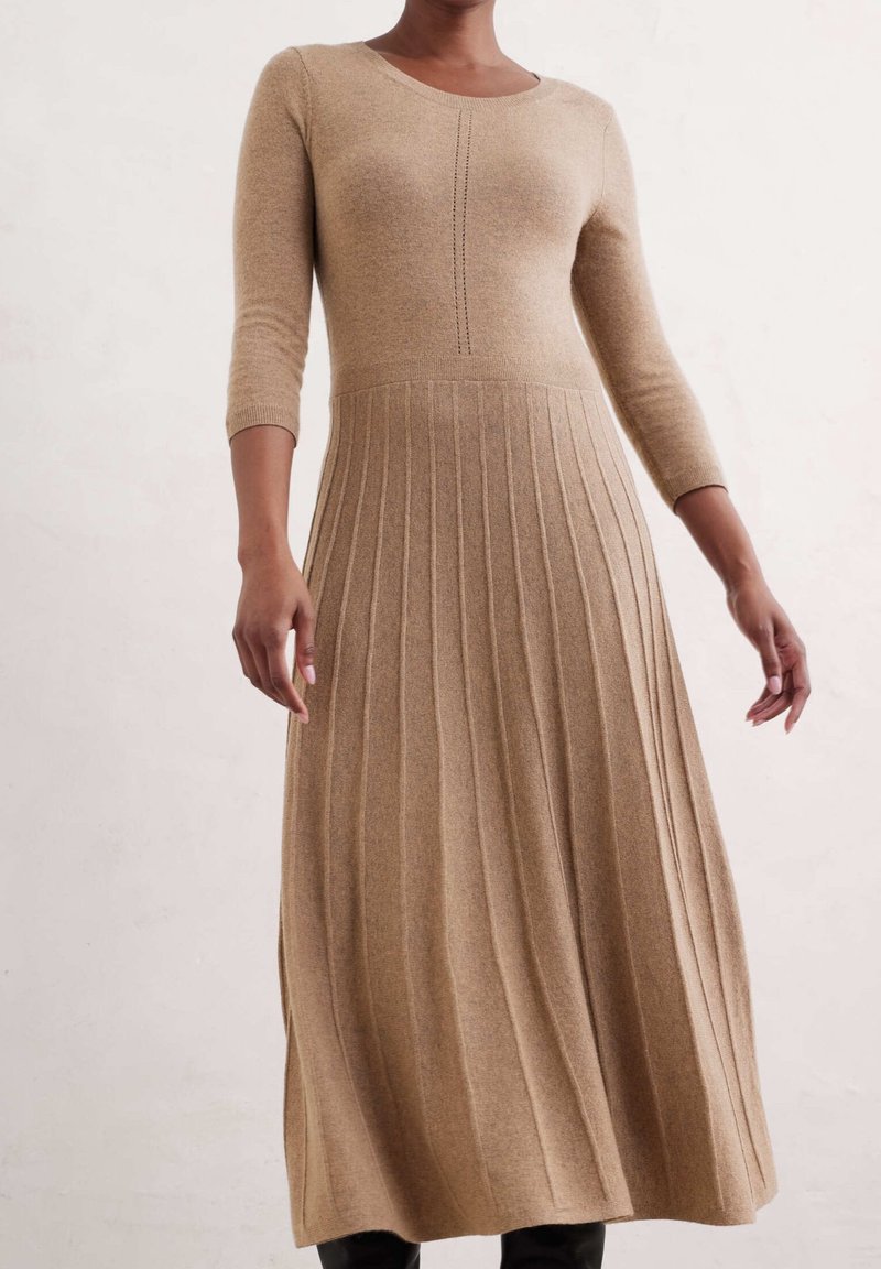 Femme portant une robe en maille beige à manches longues avec une jupe côtelée et une couture décorative au centre avant, debout devant un fond uni.