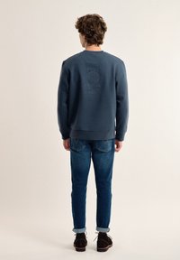 Sudadera azul marino con detalle del logo en relieve en la parte posterior, combinada con jeans de mezclilla. El conjunto tiene un ajuste casual y una textura suave.