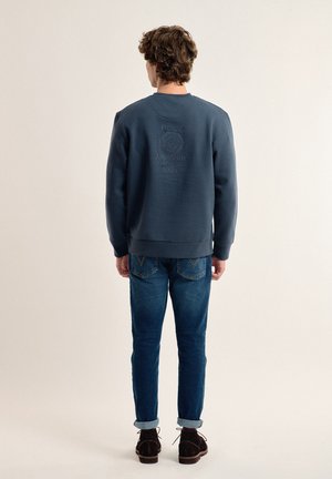 Sudadera azul marino con detalle del logo en relieve en la parte posterior, combinada con jeans de mezclilla. El conjunto tiene un ajuste casual y una textura suave.