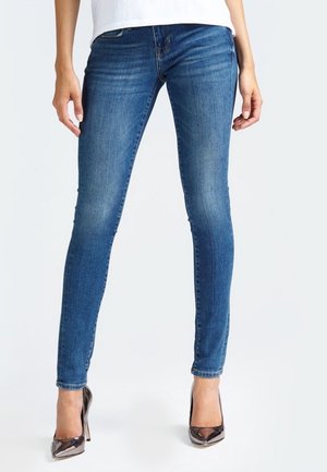 Kobieta ubrana w dopasowane niebieskie dżinsy i metaliczne, szpiczaste szpilki, stojąca na tle jednolitego białego tła.