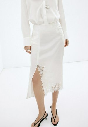 Falda midi blanca con encaje en el borde y abertura frontal, combinada con camisa blanca abotonada y sandalias negras con tiras y tacón bajo sobre fondo neutro.