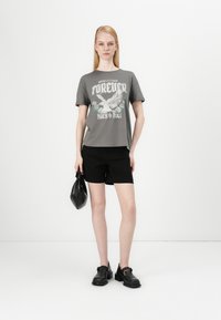 T-shirt gris avec un imprimé graphique représentant un aigle et des roses, associé à un short noir et des chaussures noires à enfiler. Le modèle tient un sac noir.
