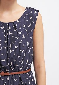 Laivastonsininen mekko, jossa on valkoinen linnunpainatus, pyöreä kaula-aukko, laskokset ja ruskea nahkavyö. Olkapäissä koristeelliset napit. Liukas kangas.