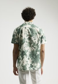 Camisa de manga corta en color crema con estampado floral tropical en verde. Presenta un cuello de tipo camp y un corte relajado. Tejido ligero con textura suave.