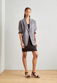 Blazer de pana gris con mangas enrolladas, botones de doble botonadura, llevado sobre un vestido negro ajustado. Sandalias de cuña negra con tiras en el tobillo.