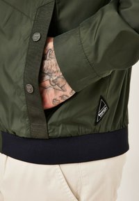 Olivgrüne Jacke mit glatter Textur, ausgestattet mit einem gerippten marineblauen Bund, Knopfverschlüssen und einem schwarzen dreieckigen Logo-Patch.