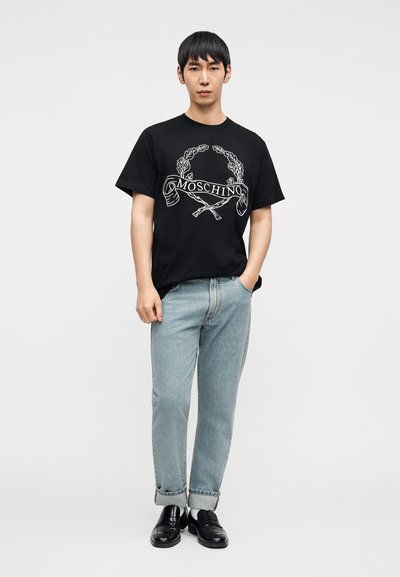 Μαύρο μπλουζάκι με λευκό λογότυπο Moschino και διακοσμητικά στοιχεία. Συνδυασμένο με ανοιχτό μπλε τζιν με ρεβέρ και μαύρα παπούτσια.