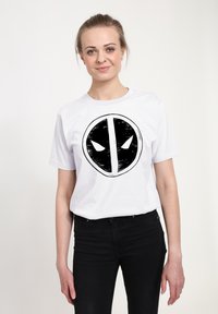Vit bomullströja med en djärv svart grafik av ett ansikte med stora ögonöppningar. Stylerad med korta ärmar och rund halsringning.