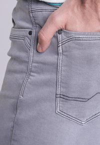 Jeans de mezclilla gris con corte ajustado, que presentan costuras en contraste, detalles en los bolsillos delanteros y una textura suave. Mano posicionada en el bolsillo.