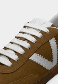 Καφέ σουέντ αθλητικό παπούτσι με λευκά κορδόνια, που διαθέτει μεγάλο λευκό λογότυπο και υφασμάτινες λεπτομέρειες στο πλάι. Λείο καουτσούκ πάτο και μαλακό άνω μέρος.