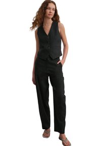 Gilet nero con chiusura a bottoni e due tasche frontali, abbinato a pantaloni neri larghi. Modello con sandali argento.