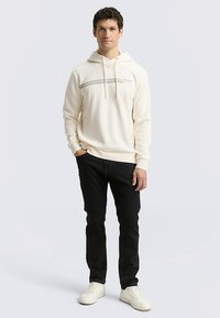 Roomkleurige katoenen hoodie met een voorzak en grijze accentstrepen, gecombineerd met zwarte jeans en witte sneakers; staande figuur, neutrale achtergrond.