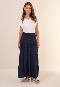 Gonna maxi blu scura con design a strati, vita elasticizzata arricciata, abbinata a una maglietta bianca a scollo a V e sandali metallici.