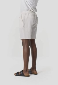 Persona que lleva pantalones cortos blancos con textura y sandalias negras de doble tira, de pie contra un fondo claro y liso.