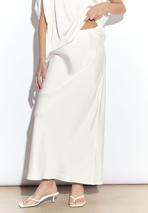 Kobieta w białej szmizjerce z satynowej maxi spódnicy, białych sandałkach na niskim obcasie z paskami, unosząca biały top, aby odsłonić pasek spódnicy.