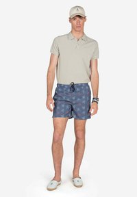 Camisa polo beige, pantalones cortos de baño azules con patrones náuticos rojos y blancos, y gorra beige; calzado casual con una textura ligera.