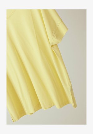 Vaaleankeltainen lyhythihainen puuvillainen t-paita pyöreällä pääntiellä ja sivuhalkioilla, yksivärisellä vaalealla taustalla.