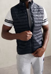 Muž v bílém tričku s černými pruhy a světle šedých teplácích si zapíná černou bezrukávovou péřovou vestu.