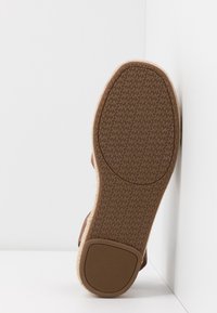 Semelle d'une sandale marron avec un motif "MK" texturé au fond, placée debout contre un mur blanc.