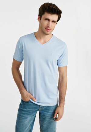 Jasnoniebieska koszulka z dekoltem w serek, wykonana z miękkiego materiału, z krótkimi rękawami i małym haftowanym logo na piersi. W zestawieniu z dżinsowymi szortami.