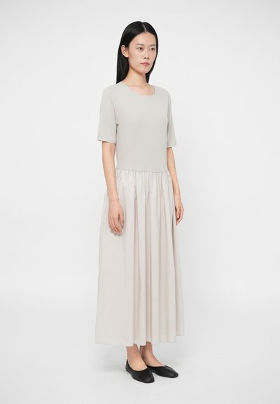 Šviesiai pilkas iki kelių ilgio suknelė su prigludusiu viršumi, trumpomis rankovėmis ir raukšlėta sijonu. Apsirengta juodais plokščiais bateliais. Drabužis pagamintas iš lygios, minkštos medžiagos.