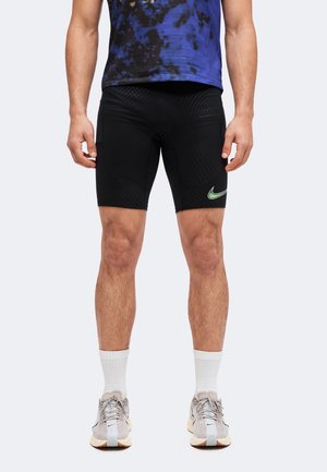 Άνδρας που φοράει μαύρα συμπιεστικά σορτς Nike, λευκές κάλτσες μέχρι τον αστράγαλο, ανοιχτό γκρι αθλητικά παπούτσια Nike και μπλε-μαύρο πουκάμισο με σχέδιο.