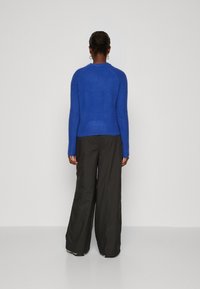 Pull bleu en tricot avec une coupe ajustée, manches longues et poignets côtelés, associé à un pantalon noir ample.