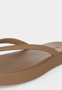 Κοντινό πλάνο ενός καφέ σαγιονάρα Crocs, που δείχνει υφασμένο πέλμα και λεπτά λουράκια με το λογότυπο Crocs στο πλάι.