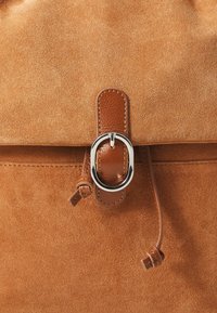 Brown suede backpack with a smooth leather flap, featuring a silver buckle closure and drawstring accent, showcasing a textured surface.