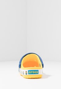 Gula Crocs med blå rem, med ett fantasifullt mönster av minions längs sidan. Lättviktsmaterial och rundad form. Öppen bakdesign.