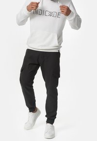 Hvid hoodie med "INDICIDE" tekst, kengurulomme; parret med sorte cargo bukser med sidelommer; hvide sneakers fuldender looket.