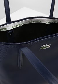 Bolso tote azul marino hecho de material texturizado con cierre de cremallera, que cuenta con un logo verde de Lacoste y una etiqueta impresa en el interior.