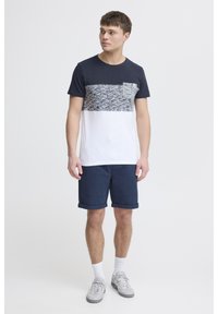 Marineblå og hvit t-skjorte med kort erme, med strukturert, mønstret brystpanel og lomme. Kombinert med marineblå shorts og grå joggesko.
