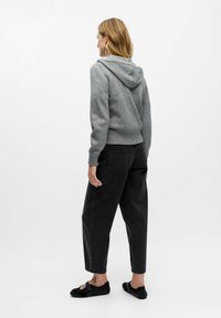 Kobieta odwrócona tyłem, ubrana w szary hoodie, luźne czarne dżinsy oraz czarne buty wsuwane, z widocznym tatuażem na kostce.