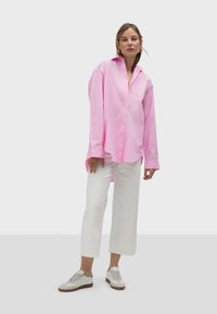 Rosa Baumwollovershirt mit langen Ärmeln, Knopfverschluss und spitzer Kragen; kombiniert mit weißen weiten Hosen und hellen Sneakern.
