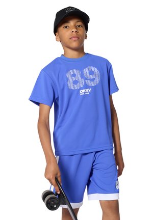 Adolescent portant une tenue bleue DKNY et une casquette noire, tenant un skateboard noir aux roues bleues dans sa main droite.