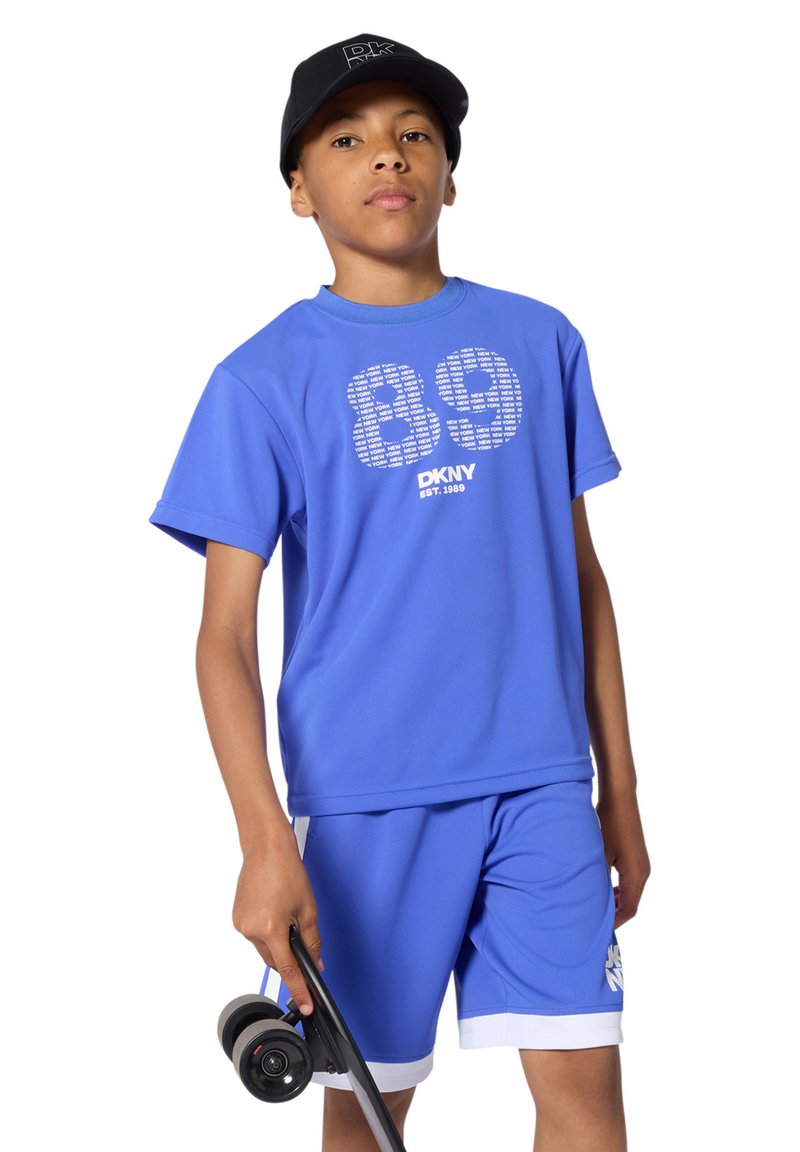 Adolescent portant une tenue bleue DKNY et une casquette noire, tenant un skateboard noir aux roues bleues dans sa main droite.