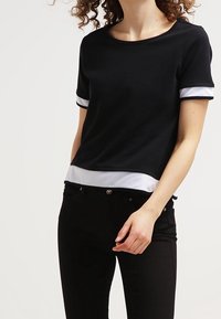 Černý top s krátkým rukávem a kulatým výstřihem, zdobený bílými pruhy na rukávech a bílým lemem. Vyrobený z hladké, elastické tkaniny.
