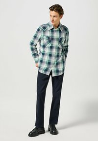 Rutigt flanellskjorta i blå, grön och vit nyanser, med spread collar, två bröstfickor och knäppning fram. Svarta byxor och skor.