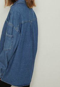 Persona che indossa una giacca di jeans blu a taglio morbido con una tasca sulla manica, mostrata di spalle su uno sfondo chiaro e semplice.