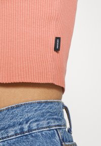 Růžový žebrovaný crop top s rovním lemem, kombinovaný s modrými denimovými šortkami s vysokým pasem, které mají na vrchu ozdobnou visačku.
