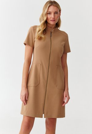 Vestido de manga corta, color beige, con cremallera frontal y dos grandes bolsillos de parche, que presenta un cuello alto y una textura de tela suave.