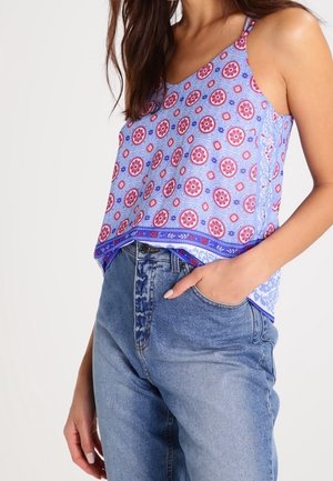 Vrouw die een mouwloze blauw met patroon blouse draagt, ingestopt in een lichtblauwe, hooggetailleerde spijkerbroek, met één hand in een zak.