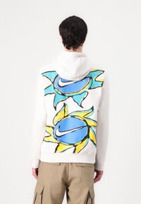 Huvtröja i vitt tyg med en stor blå och gul solgrafik på baksidan, med svarta konturer och Nike-logotypdetalj.