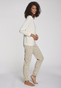 Jonge vrouw die glimlacht, gekleed in een crème kleurige lange mouw top en lichtgekleurde pyjamabroek met een rood suikerrietpatroon, staande op blote voeten.
