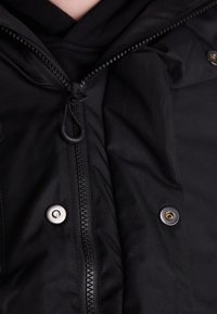 Chaqueta negra con cremallera frontal, botones a presión y un tejido texturizado. El tirador de la cremallera tiene un agarre de goma para facilitar su uso.