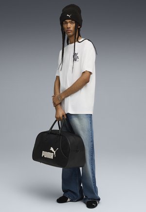 Svart duffelbag med vitt "PUMA"-logo, hålls av en person som bär en vit T-shirt, blå jeans och en svart stickad mössa.